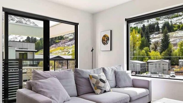 Ein modernes Wohnzimmer mit einer grauen Couch, einem Couchtisch und großen Fenstern mit Blick auf eine Berglandschaft.