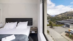 Ein Hotelzimmer mit Bett, Nachttisch, Lampe und einem großen Fenster mit Blick auf einen Parkplatz und Berge.