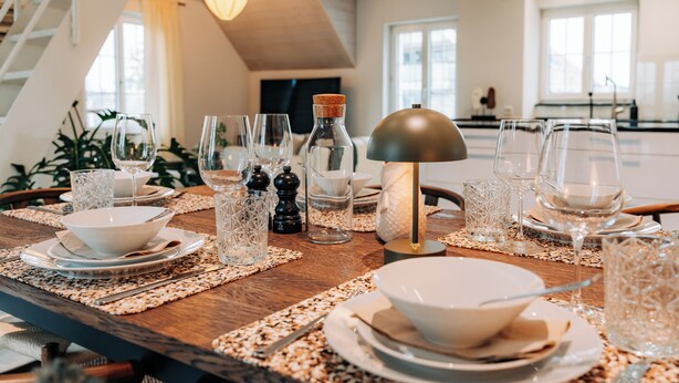 Une table à manger dressée avec de la verrerie, des assiettes et un centre de table composé d’une bougie, dans un intérieur moderne baigné de lumière naturelle.