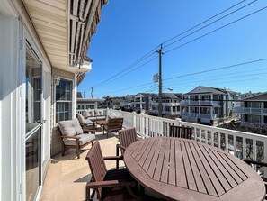 En balkon med et træbord og stole, et hvidt gelænder og udsigt til omkringliggende huse.