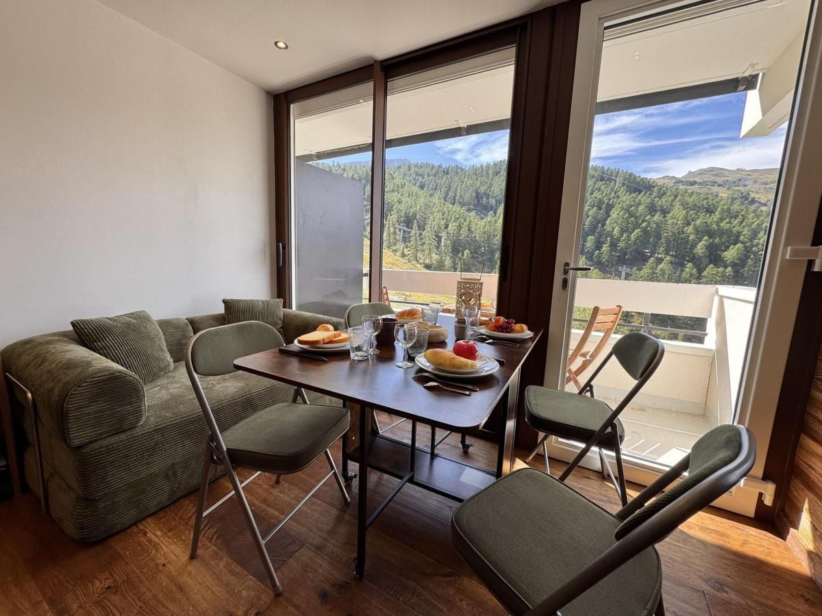A dining area with a table set for two, a sofa, and a view of a forested landscape through large windows.