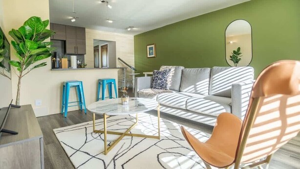 Una sala de estar moderna con un sofá, una mesa de centro y sillones. Se observa una cocina con armarios y una encimera.