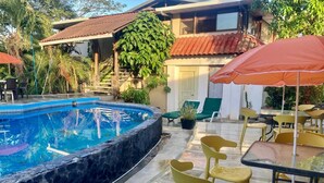 En swimmingpool med en kant av sten, omgiven av utomhusmöbler och en byggnad med ett tak täckt av tegelpannor.