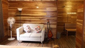 A cozy room with a wooden wall, a white sofa with colorful cushions, a standing fan, a guitar, and a bench.