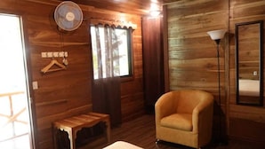 A wooden room with a bed, a chair, a small table, a fan, and a window with curtains.