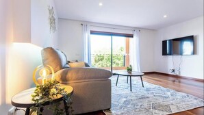 A modern living room with a sofa, a small round table, a TV mounted on the wall, and a large window with a view of greenery. - Apartamento amplo no Baía da Luz – férias perfeitas para famílias na (Lagos)