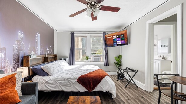 Un dormitorio moderno con un mural de paisaje urbano, una cama con manta blanca y marrón, un televisor en la pared y suelo de madera.