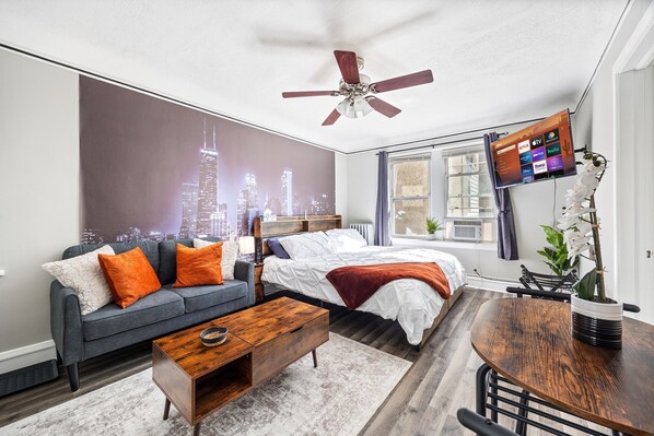 A modern bedroom with a cityscape mural, a bed with white bedding, a gray sofa with orange pillows, a wooden coffee table, and a TV mounted on the wall.