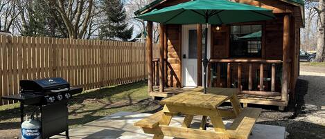Una pequeña cabaña de madera con toldo verde, una parrilla y una mesa de picnic.