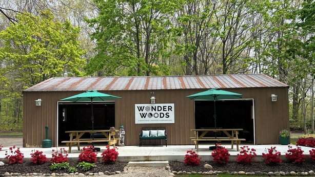 Un bâtiment rustique en bois, doté de deux portes de garage ouvertes, d’un banc et d’une enseigne sur laquelle on peut lire « Wonder Woods ».