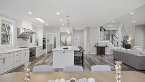 A modern kitchen with white cabinetry, a central island, and a dining area with a wooden table and chairs. - Luxe Retreat w/ Spa Sleeps 25 Designed For Groups (Charlotte)