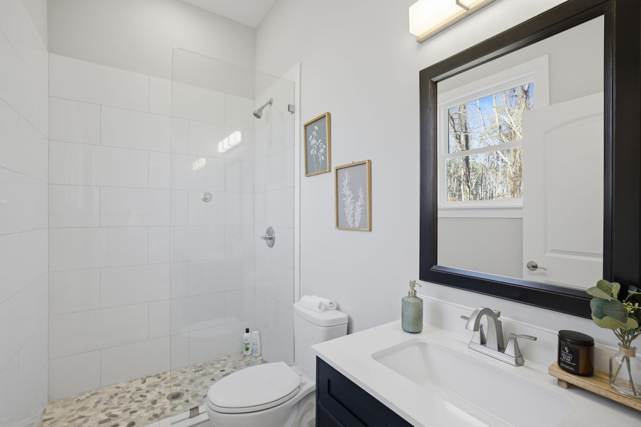 Une salle de bain moderne avec une douche en verre, des carreaux blancs, des toilettes blanches, un lavabo avec un miroir et des tableaux encadrés au mur.