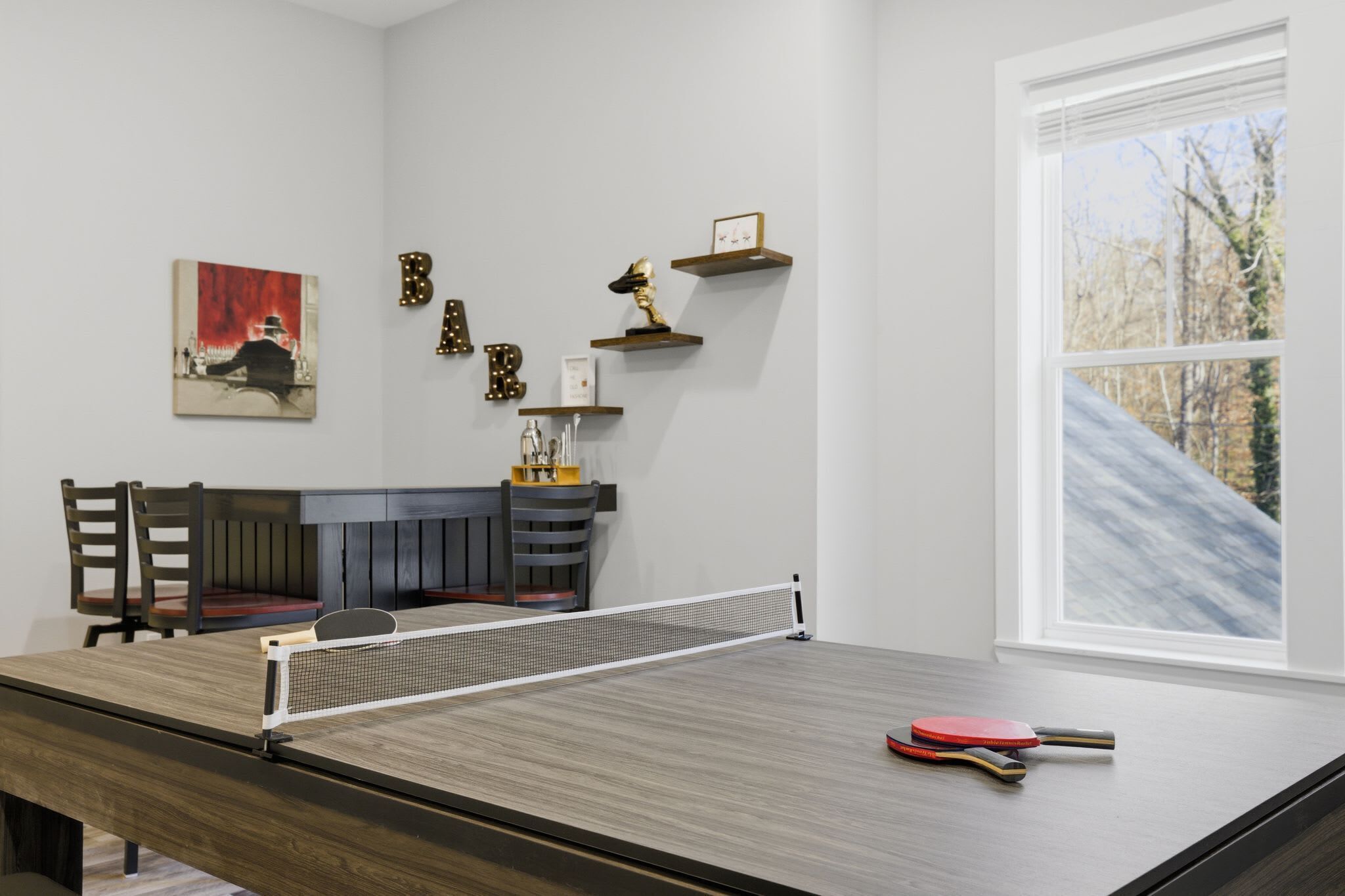 Une table de ping-pong avec un filet, deux raquettes et une balle de tennis de table. On y voit également deux chaises, un banc et un tableau accroché au mur. Des étagères ornées d’objets décoratifs sont également visibles.