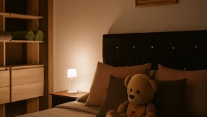 A bedroom with a bed, a teddy bear, a nightstand with a lamp, a wooden shelf unit, and a framed picture on the wall.
