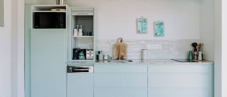 A modern kitchen with light blue cabinets, a built-in dishwasher, and a microwave. There are two hanging decorations on the wall and a wooden cutting board on the counter.