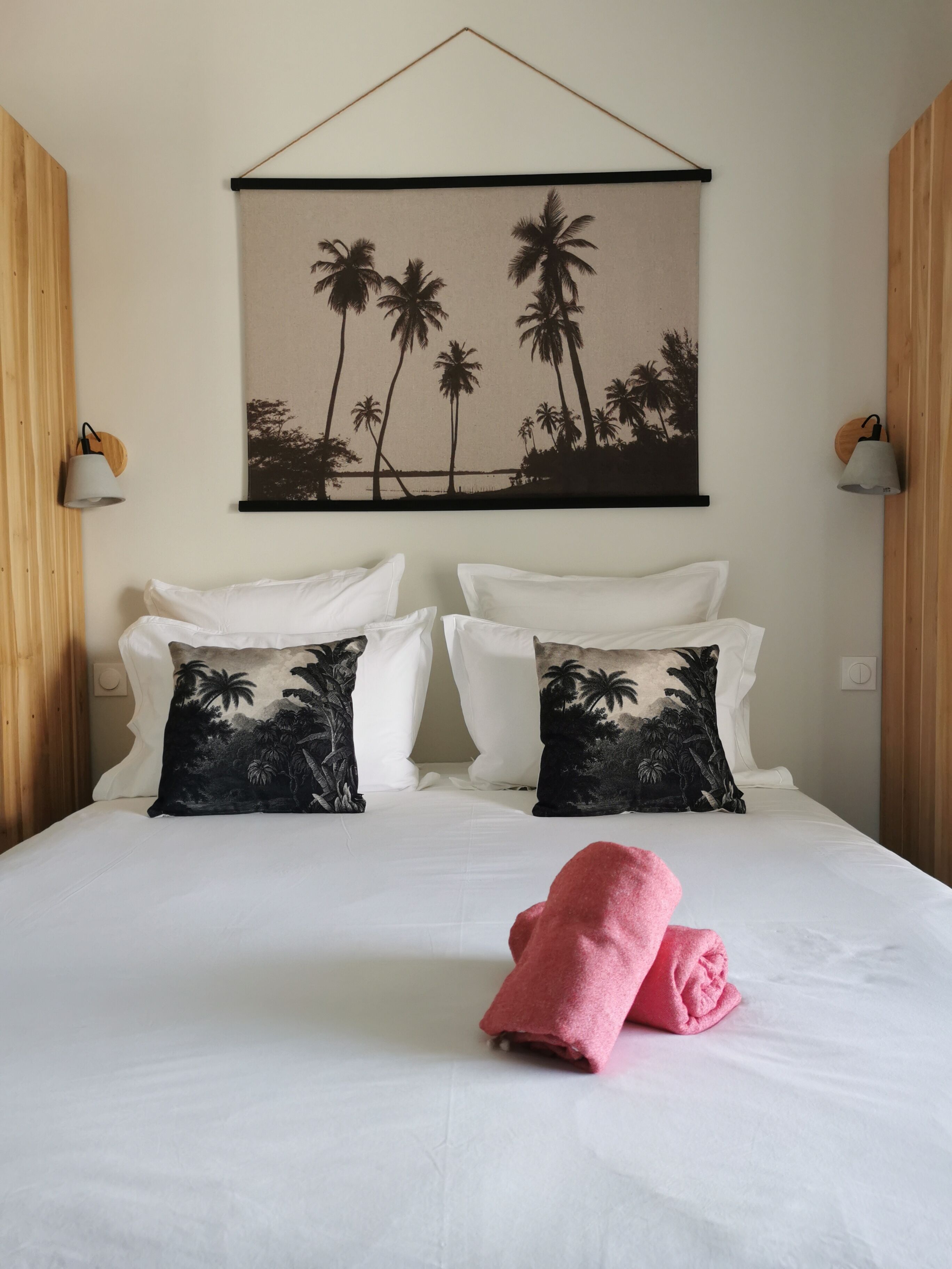A hotel room with a bed, two pillows with a palm tree design, a wall-mounted picture of palm trees, and two wall-mounted lamps.
