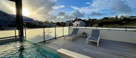 A rooftop swimming pool with lounge chairs, a wooden deck, and a view of houses in the background.