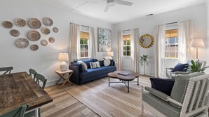 A living room with a blue sofa, a wooden coffee table, a dining table with chairs, and a ceiling fan. - Charming Cottage in Downtown Charleston (Charleston)