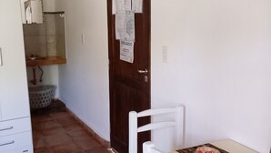 A small room with a wooden door, a white chair, a table with papers, a white cabinet, and a tiled floor. - Abril (Cafayate)