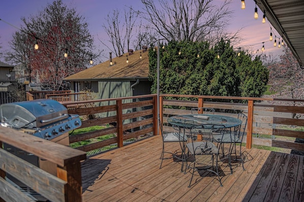 Una terraza de madera con una parrilla, una mesa de vidrio y sillas de metal, y luces colgantes.