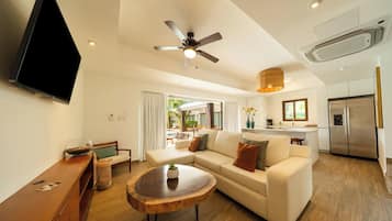 A modern living room with a sofa, coffee table, and a TV. There's a ceiling fan, a kitchen area with a refrigerator, and a view of an outdoor pool.