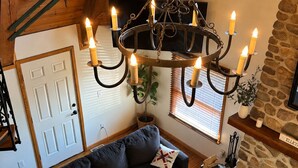 Una sala de estar acogedora con un sofá negro, una mesa de centro de madera, una araña con velas, una chimenea de piedra y un techo de madera.
