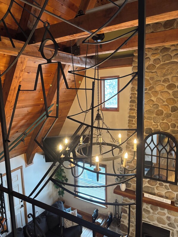 A loft-style room with exposed wooden beams, a stone wall, and a chandelier with candles. - Swiss Miss Pet-Friendly A-Frame only 6 Minutes from downtown Ellicottville! (Little Valley)