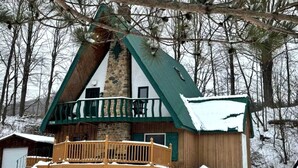 Una cabaña con chimenea de piedra, terraza de madera y techo cubierto de nieve.