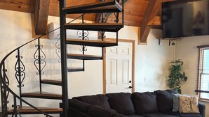 Un espacio de sala acogedor con un sofá negro, una escalera espiral de madera y un televisor de pantalla plana montado en la pared.