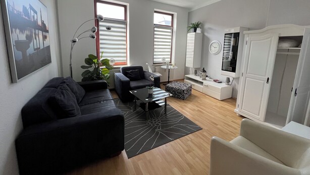 Un salon moderne avec un canapé, un fauteuil, une table basse et un tapis décoratif. Il y a deux fenêtres équipées de stores, un radiateur et un téléviseur posé sur un meuble.