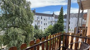 Un balcon avec des fauteuils en osier et une table, donnant sur un quartier résidentiel avec des bâtiments blancs et des arbres.