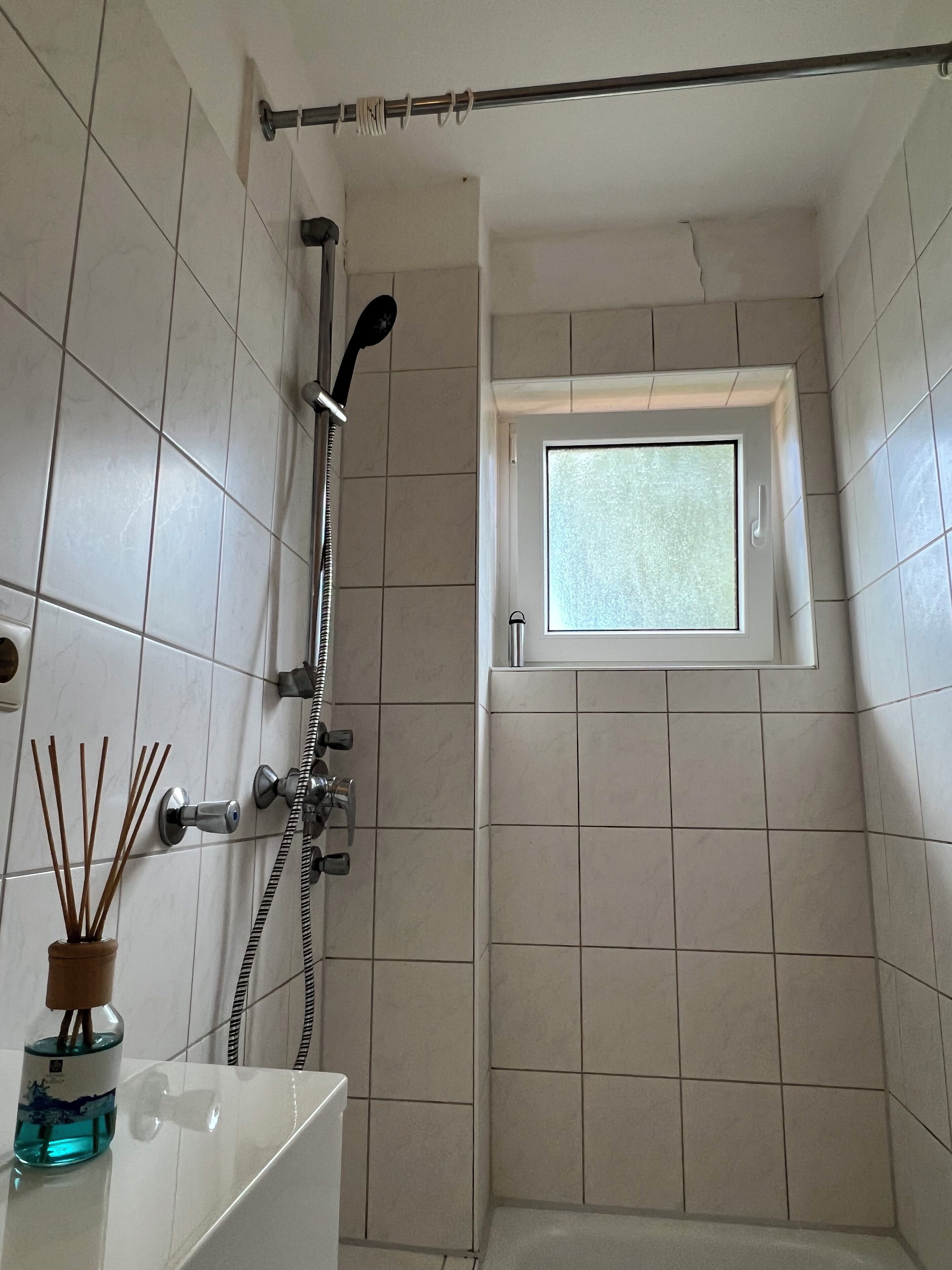 A small bathroom with a shower, a window, and a bottle of reed sticks.