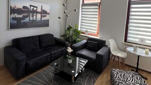 Un salon moderne avec un canapé d’angle, une table basse en verre, un pouf à motifs zébrés, une plante en pot et un tableau représentant un paysage urbain encadré.