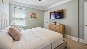 Ein Schlafzimmer mit Bett, Deckenventilator, Kommode, Fernseher und Fenster mit Jalousien.