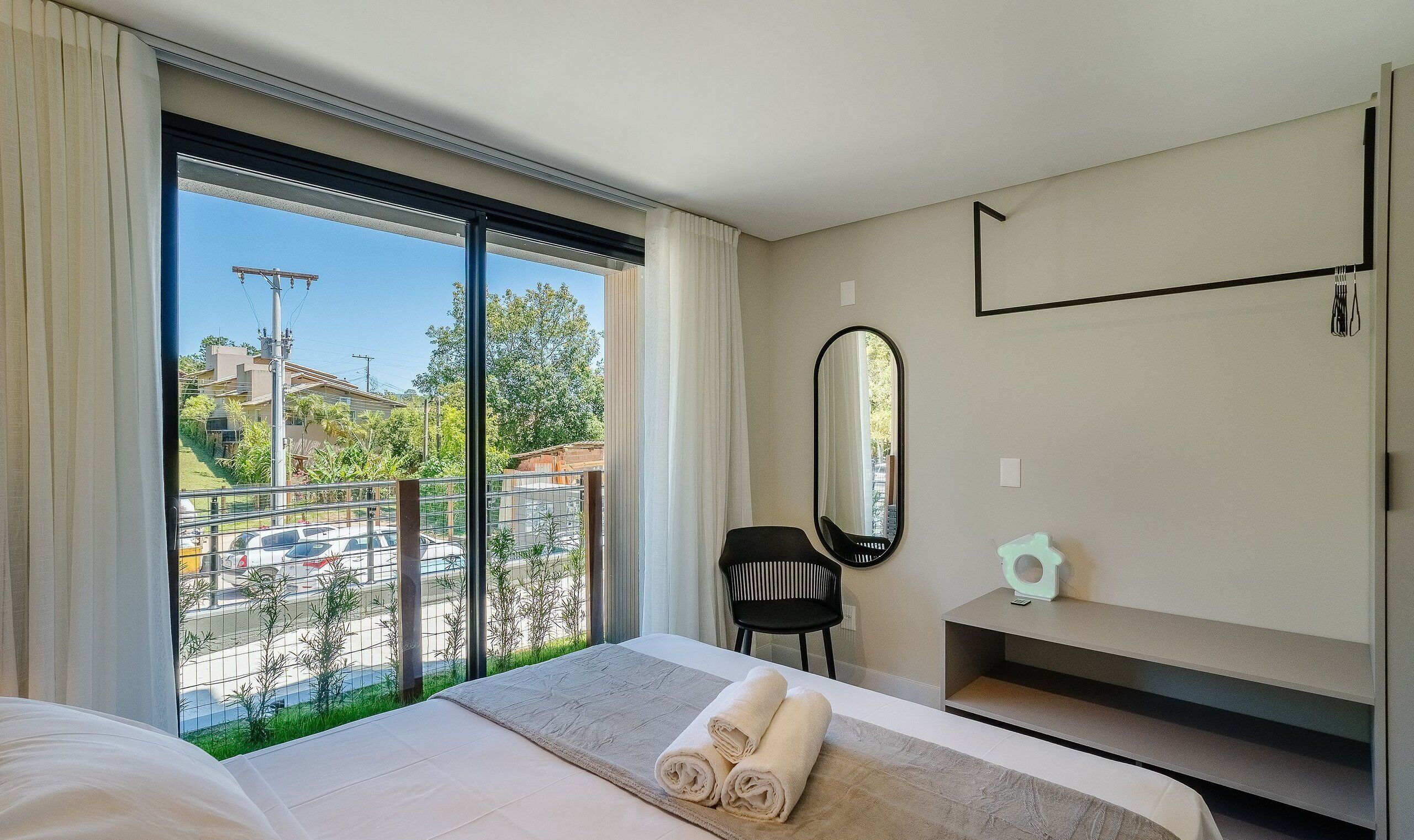 Habitación de hotel con una ventana grande, una cama, una silla, un espejo y un estante pequeño.