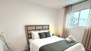 Een moderne slaapkamer met een houten vloer, een bed met wit en donker beddengoed, een nachtkastje met een lamp en een raam met gordijnen.
