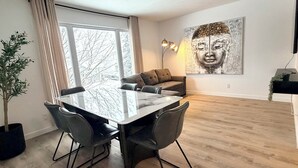 Een moderne woonkamer met een eethoek, een groot raam, een schilderij aan de muur en een flatscreen televisie.