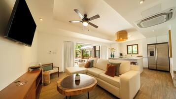 A modern living room with a sofa, coffee table, and a TV. There's a ceiling fan, a kitchen area with a refrigerator, and a view of an outdoor pool.