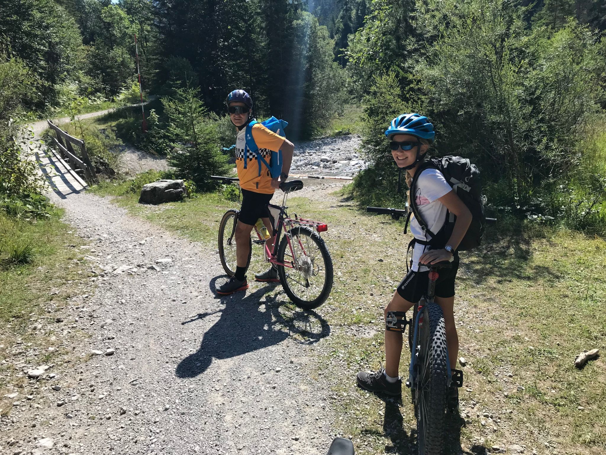 Two people on a gravel path with bicycles, surrounded by greenery and a wooden bridge.
