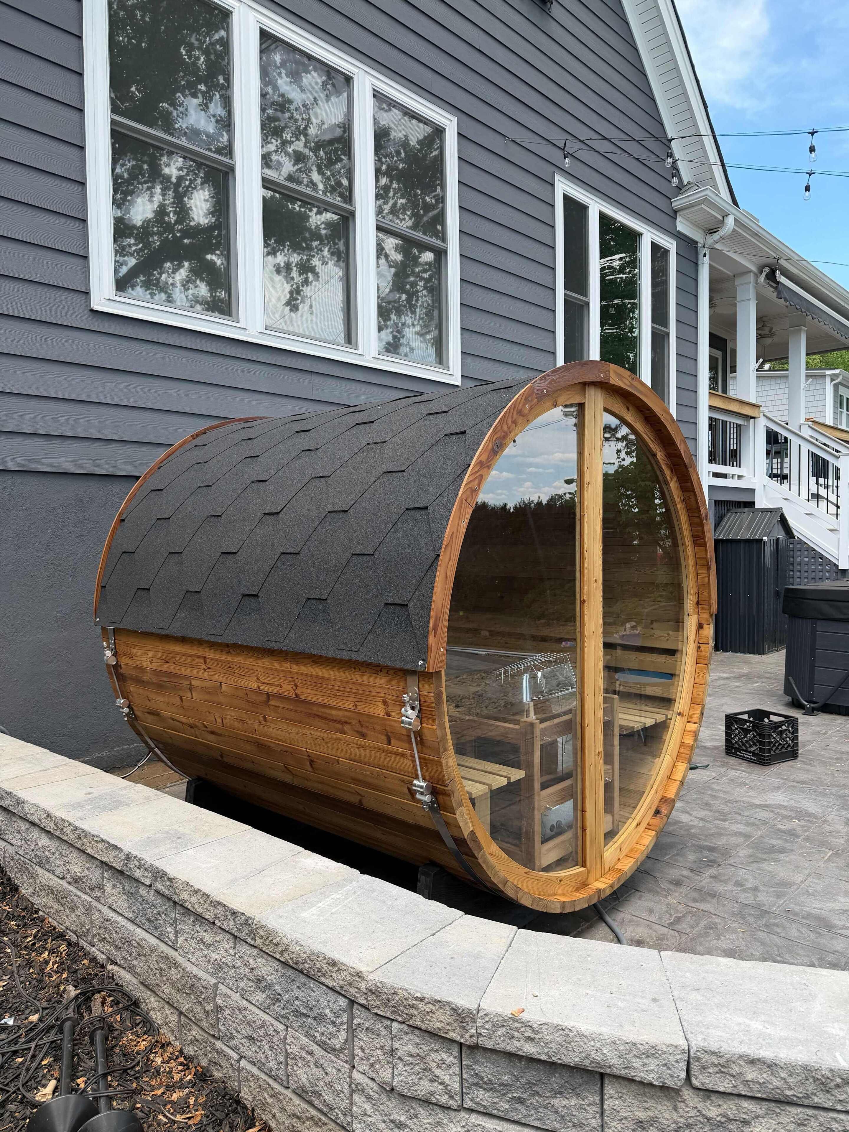 Een houten, vatvormige constructie met een glazen deur, vastgemaakt aan een huis met grijze bekleding en witte details.