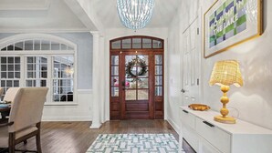 A hallway with a wooden door, a chandelier, a patterned rug, and a white console table with a lamp. - New! Lakefront-Sleeps 14-Hot Tub-Putting Green (Denver)