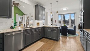 A modern kitchen with dark cabinets, a central island, and a dining area with blue chairs. - New! Lakefront-Sleeps 14-Hot Tub-Putting Green (Denver)
