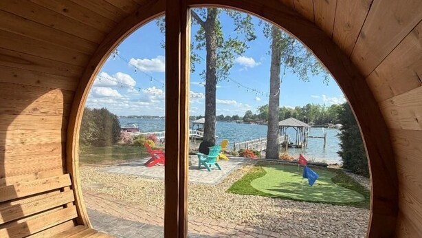 A circular wooden structure with benches inside, a glass door leading to an outdoor area with a lawn, chairs, and a view of a lake. - New! Lakefront-Sleeps 14-Hot Tub-Putting Green (Denver)