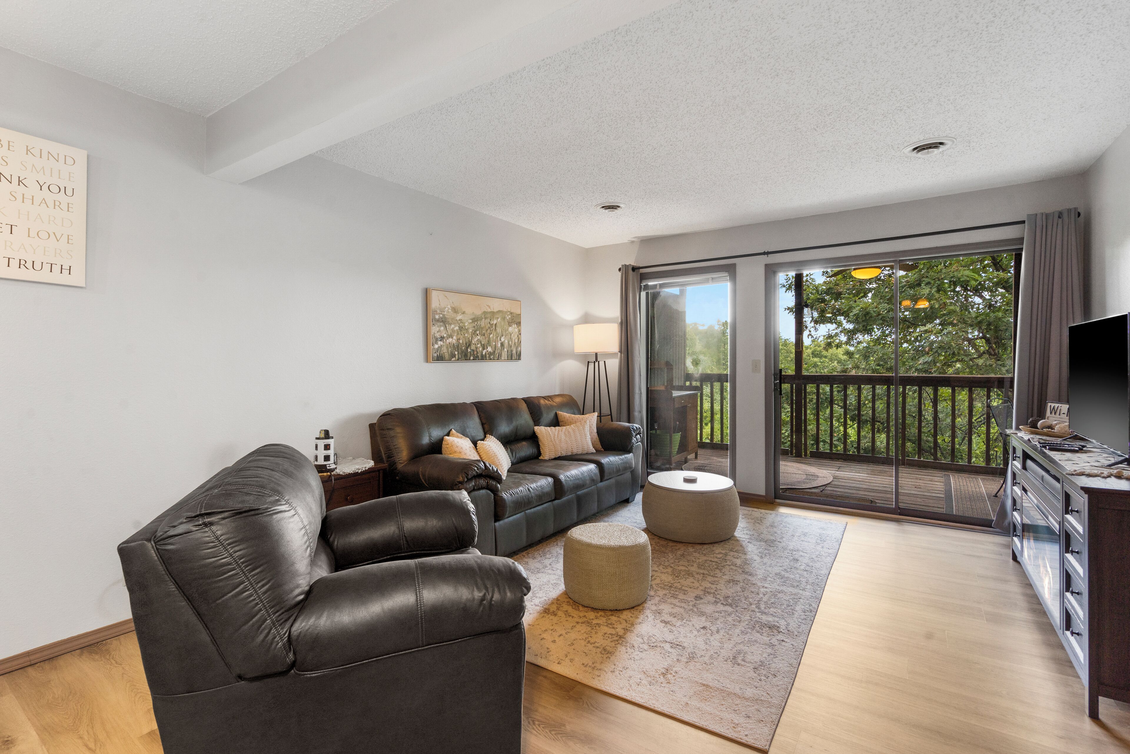 A modern living room with a leather sofa, a coffee table, and a flat-screen TV. There is a balcony with a view of trees.