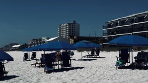 Ein Strand mit blauen Sonnenschirmen und Liegestühlen, ein Gebäude mit Balkonen und einem klaren Himmel.