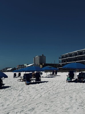 A beach with blue umbrellas and beach chairs, a building with balconies, and a clear sky. - Beach Resort 5th floor, zero depth pool, steps to the beach! (Miramar Beach)