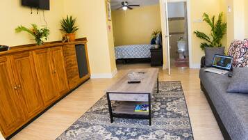 A living room with a sofa, coffee table, TV, and a small kitchen area with cabinets and a refrigerator.