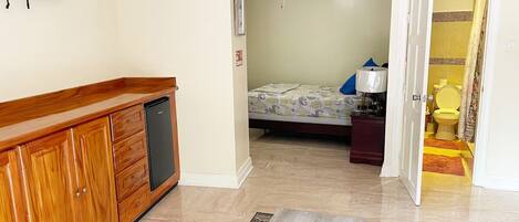 Une chambre d’hôtel compacte, dotée d’un meuble en bois, d’un ventilateur de plafond, d’un lit recouvert d’une courtepointe à motifs, d’une petite table de chevet avec une lampe, et d’une salle de bain visible à travers une porte ouverte.