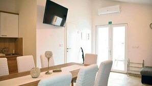 A hotel room with a wooden dining table, white upholstered chairs, a flat-screen TV mounted on the wall, a small kitchen area with a sink, and a door leading outside. - Amazing apartment in Vrsi with WiFi (Vrsi)