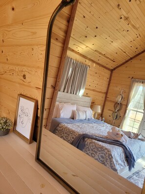 A wooden cabin bedroom with a bed, curtains, a mirror, and a framed picture on the wall. - Bohío Retreat - Cabin and Creek View on 42 Acres (Steelville)